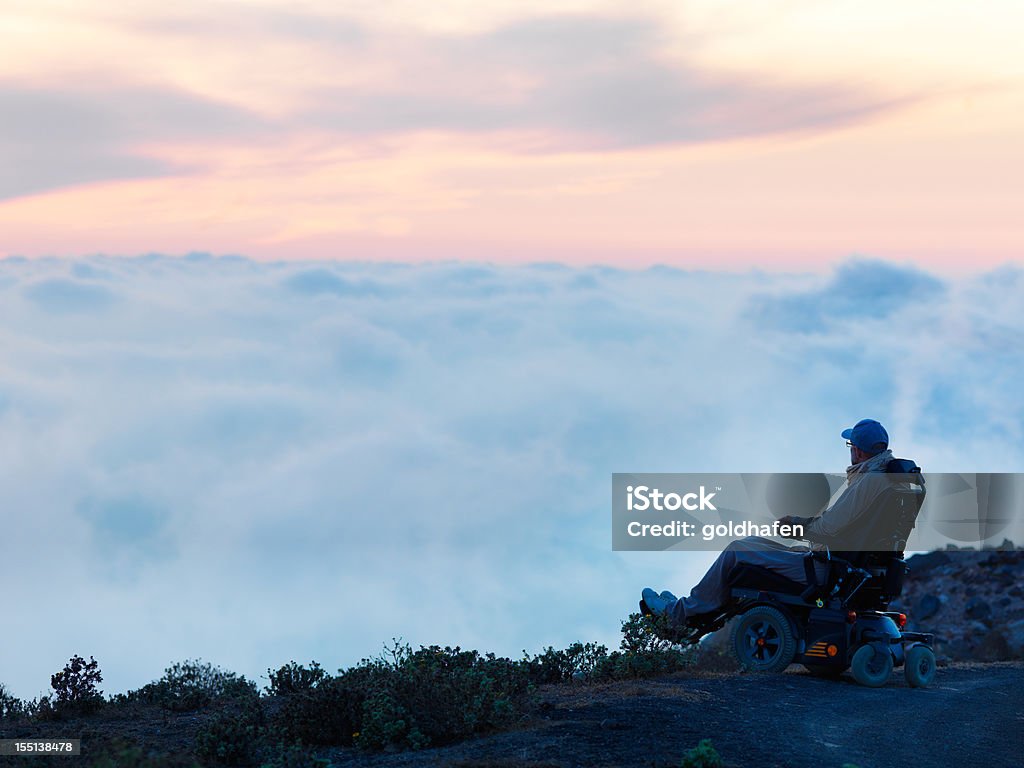 A man in a power wheelchair pauses at the edge of a mountain road, looking out over a sea of clouds at dusk