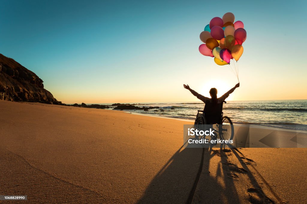 A silhouetted person in a wheelchair on an empty beach at sunset, holding a bunch of colorful balloons overhead