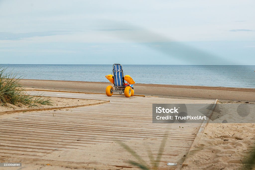 An empty beach wheelchair with yellow balloon wheels waits on a boardwalk facing a calm sea