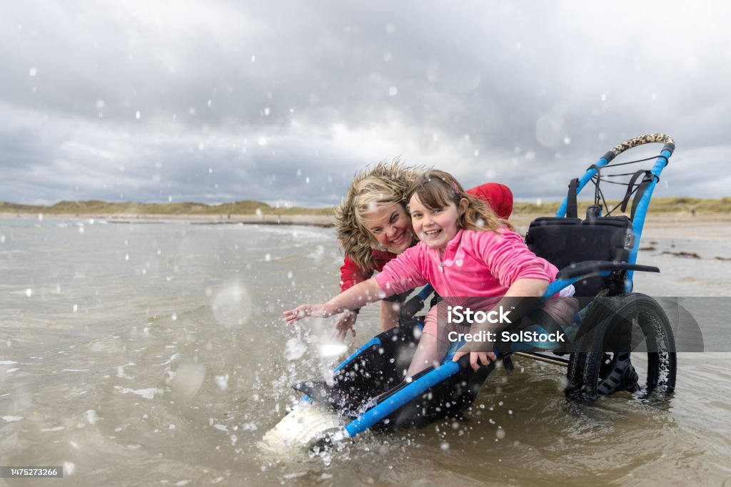 A young girl in a blue beach wheelchair laughs as a wave splashes around her, her mother beside her in the shallow water
