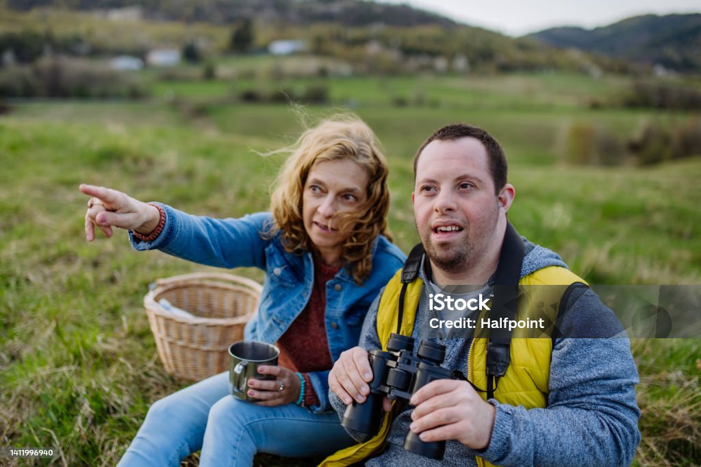 A young man with Down syndrome holds binoculars while his companion points to something across the meadow