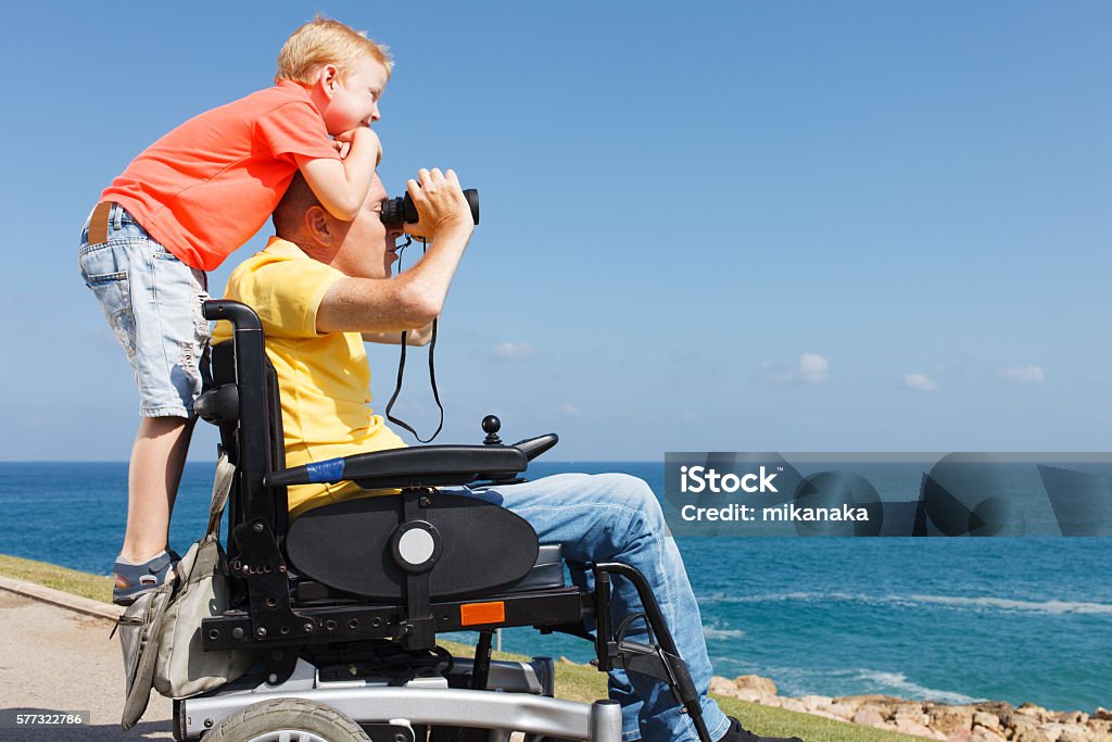 A father in a power wheelchair watches the sea through binoculars while his young son leans on his shoulders