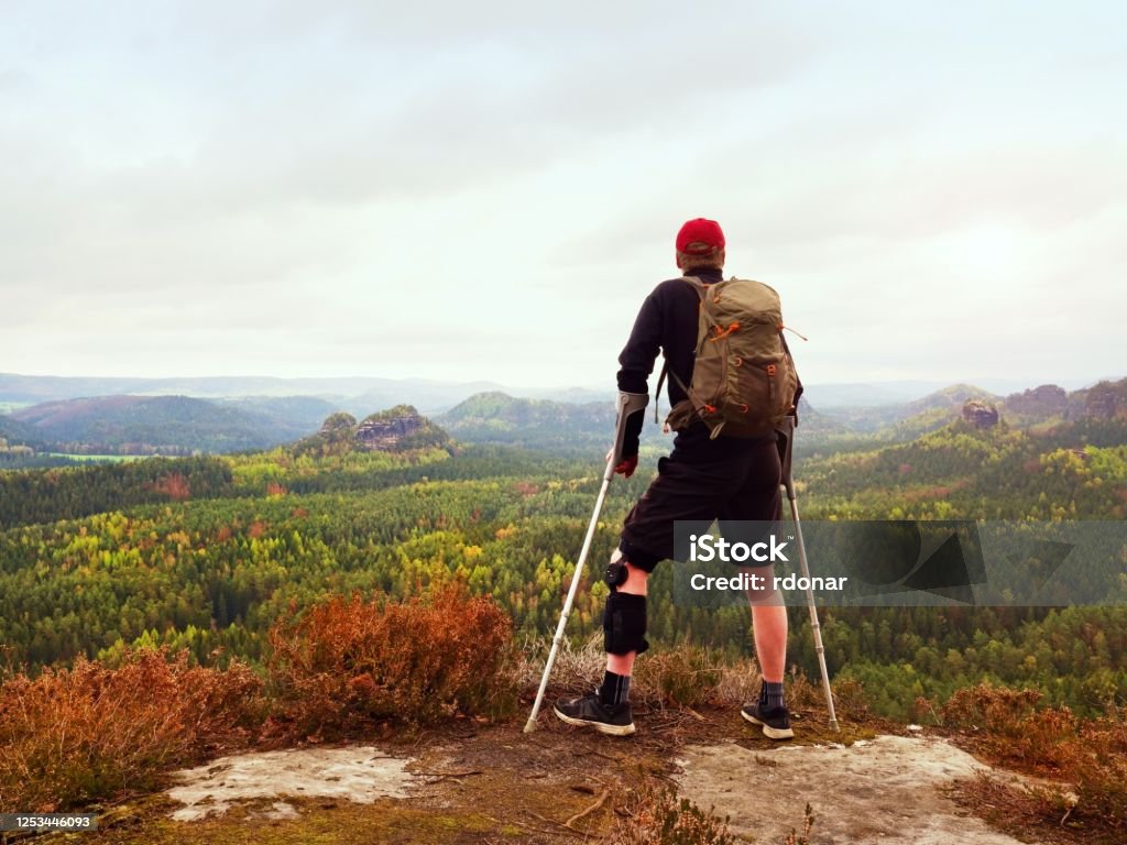 A hiker with forearm crutches and a leg brace stands at a rocky overlook above a forested valley