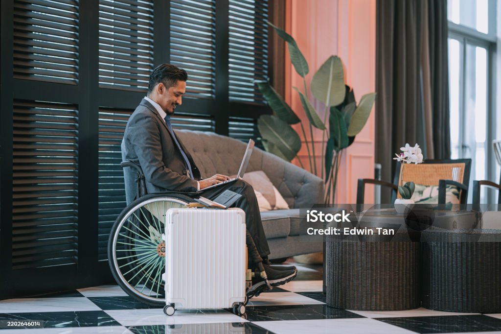 A businessman in a wheelchair works on his laptop in an elegant hotel lobby, suitcase at his side