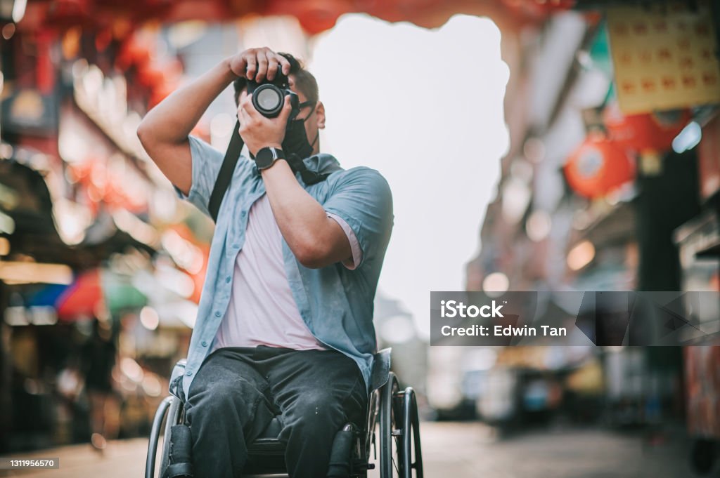 A man in a wheelchair raises a camera to photograph a lantern-lined market street