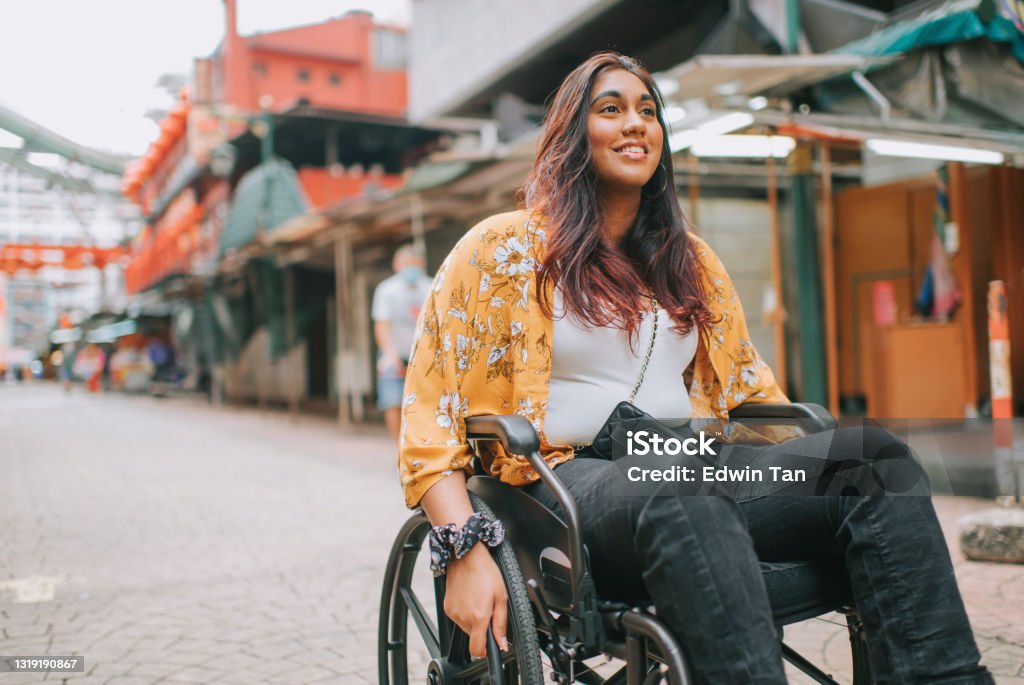 A smiling woman in a wheelchair explores a lively old market street, looking up at the stalls around her