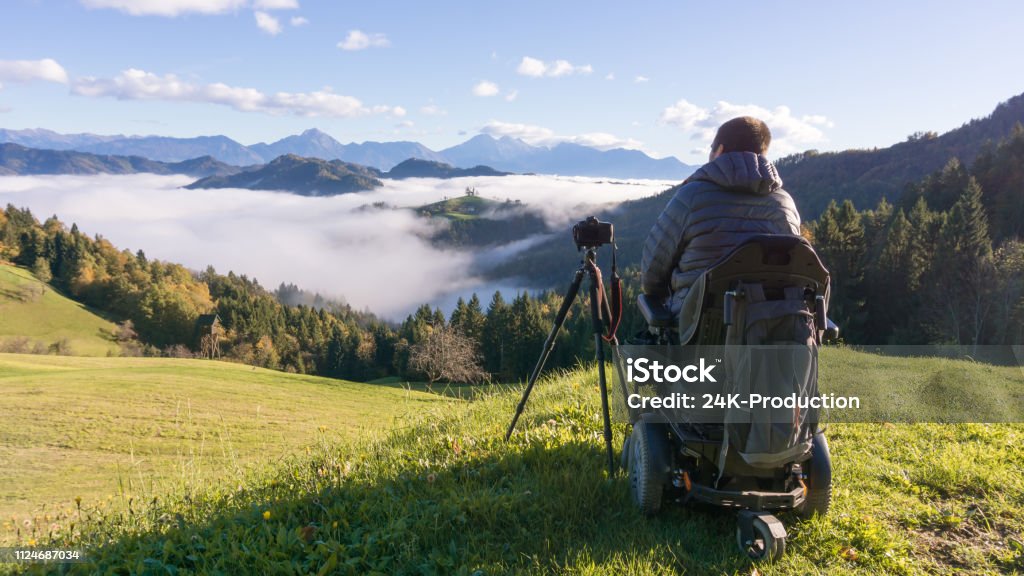 A man in a power wheelchair on a green hillside, camera on a tripod beside him, overlooking a valley of morning fog