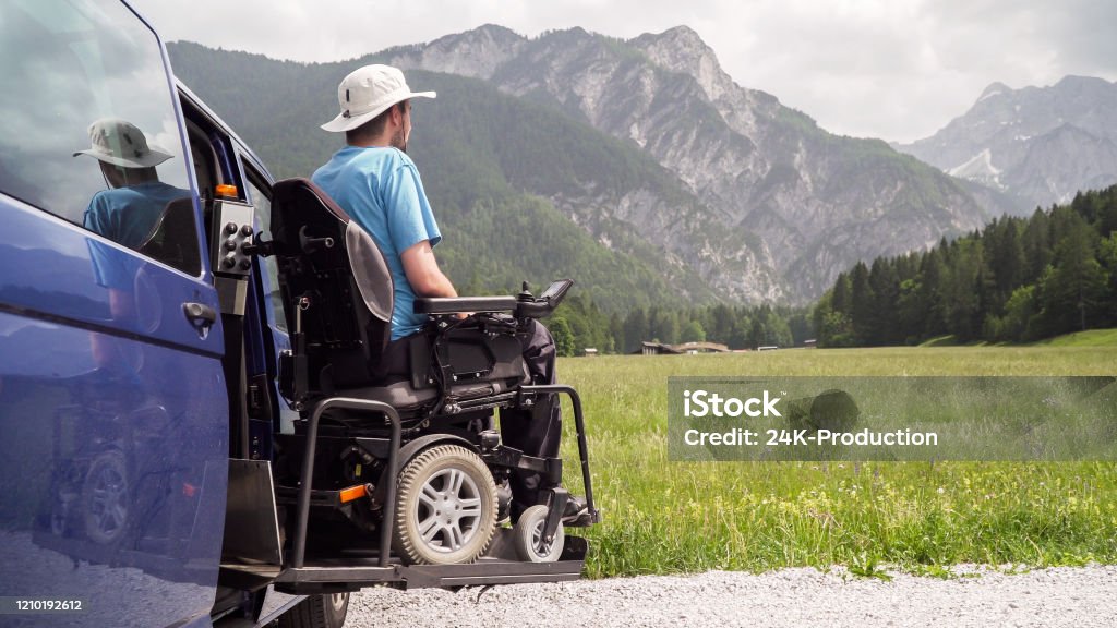A man in a power wheelchair on his van's lift platform, facing an alpine meadow and grey mountain peaks