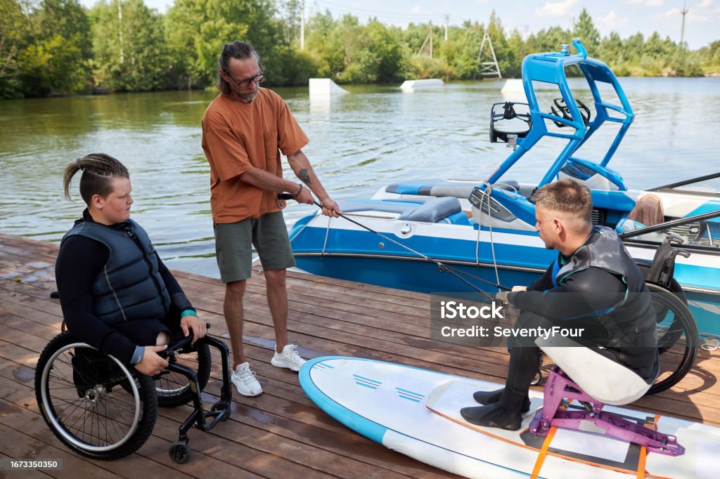At a lakeside dock, a young wheelchair user watches a friend on an adaptive wakeboard rig prepare to ride