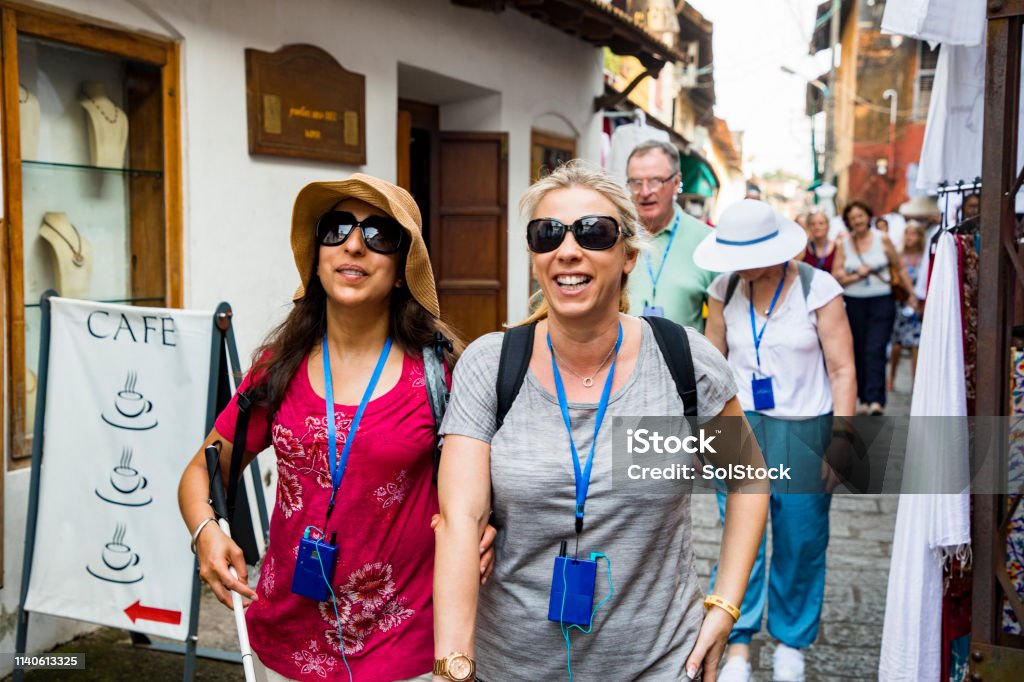 Two women walk arm in arm through an old town lane, one using a white cane, both smiling in sun hats