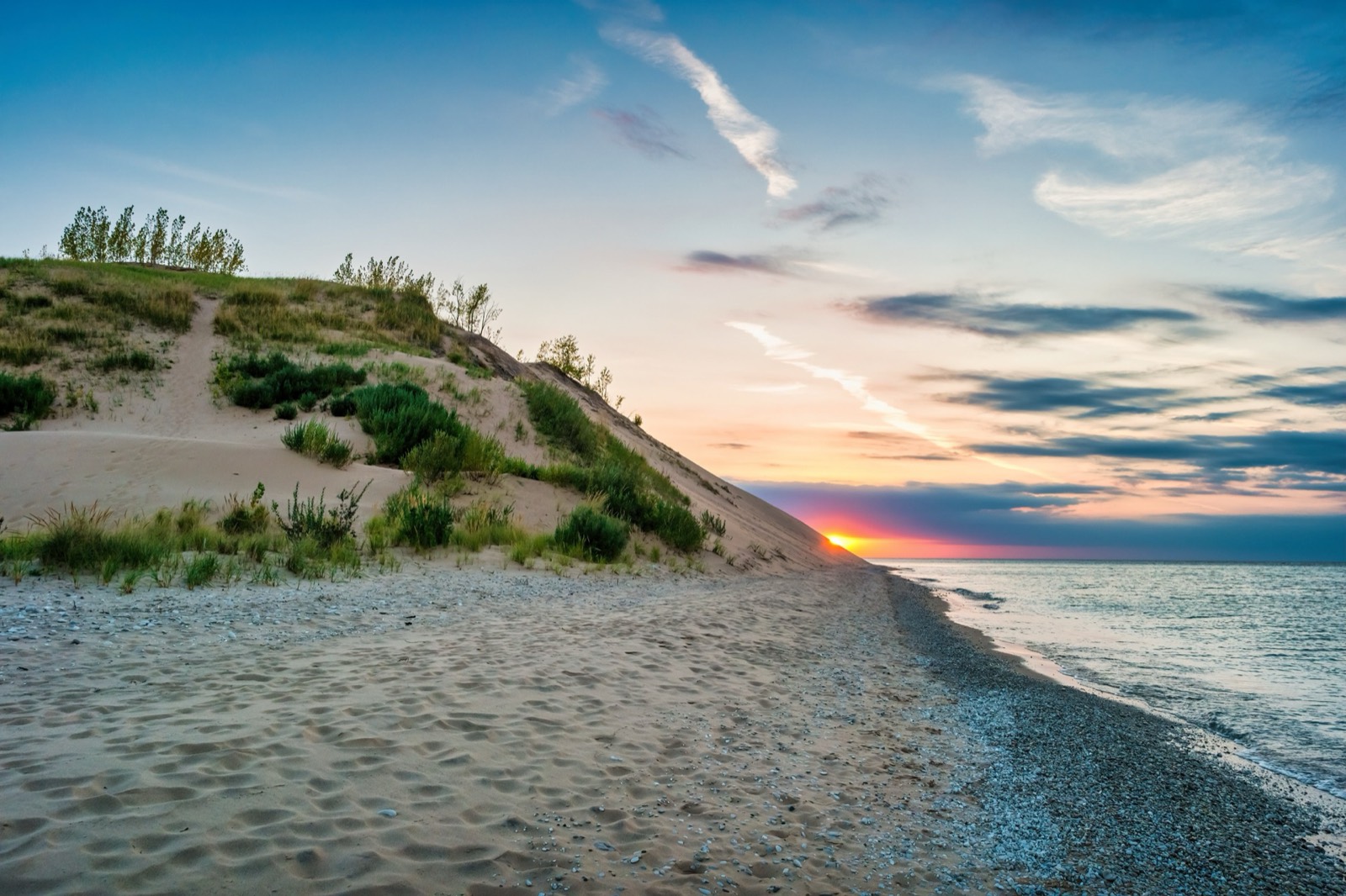 A quiet sandy beach at golden hour, a grassy dune rising beside calm water