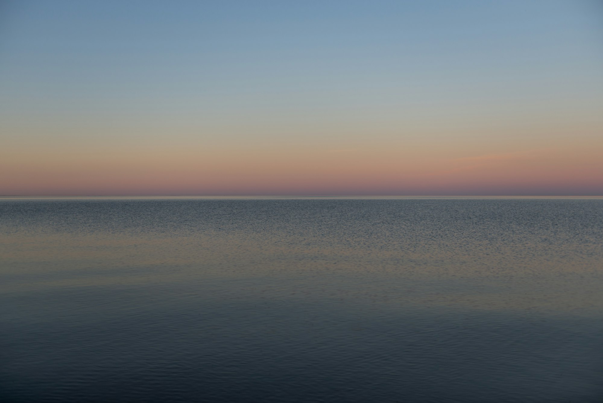 A perfectly calm sea at dusk under a soft blue and peach sky, the horizon a clean level line